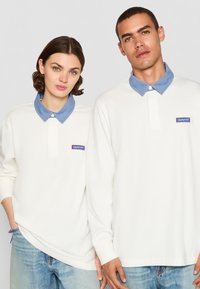 Camisas polo blancas de manga larga con cuellos de mezclilla, con un parche del logo de GANT. Tejido suave y corte relajado, llevadas por dos modelos.