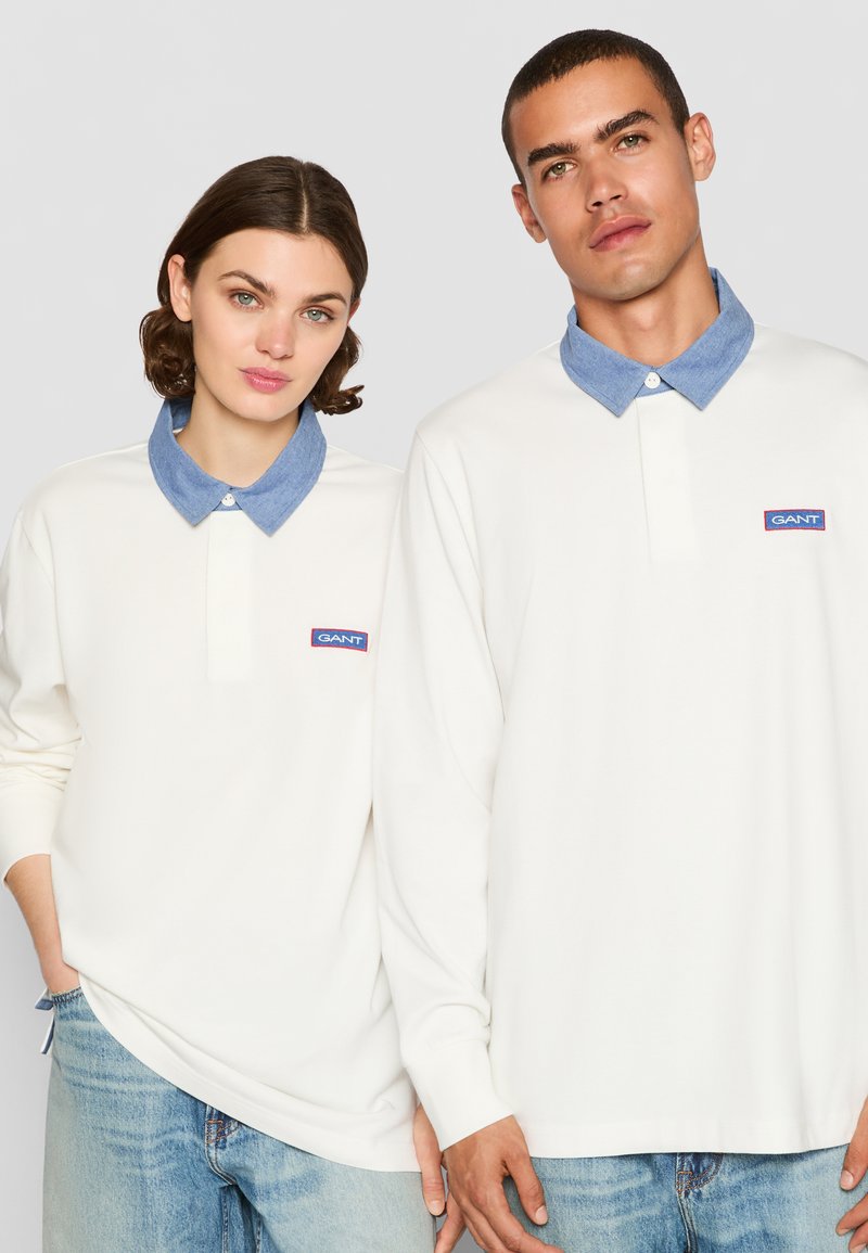Weiße Langarm-Polohemden mit Denim-Kragen, ausgestattet mit einem GANT-Logo-Patch. Glattes Material und lockere Passform, getragen von zwei Modellen.