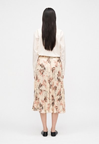 Woman with long dark hair stands facing away, wearing a white blouse, floral pleated midi skirt, and black flat shoes on a plain background.