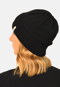 Black knitted beanie with a snug fit, featuring a folded cuff and a small white brand tag on the side, textured with a ribbed pattern.