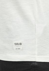 Hvid kortærmet t-shirt med en struktureret finish, der har et rektangulært mærke med "SOLID" og "DENMARK" trykt i sort.