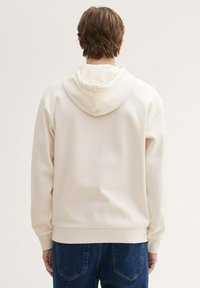 Felpa beige chiara con un cappuccio ampio, polsini e orlo a coste, e una trama di tessuto liscia, abbinata a jeans denim blu.