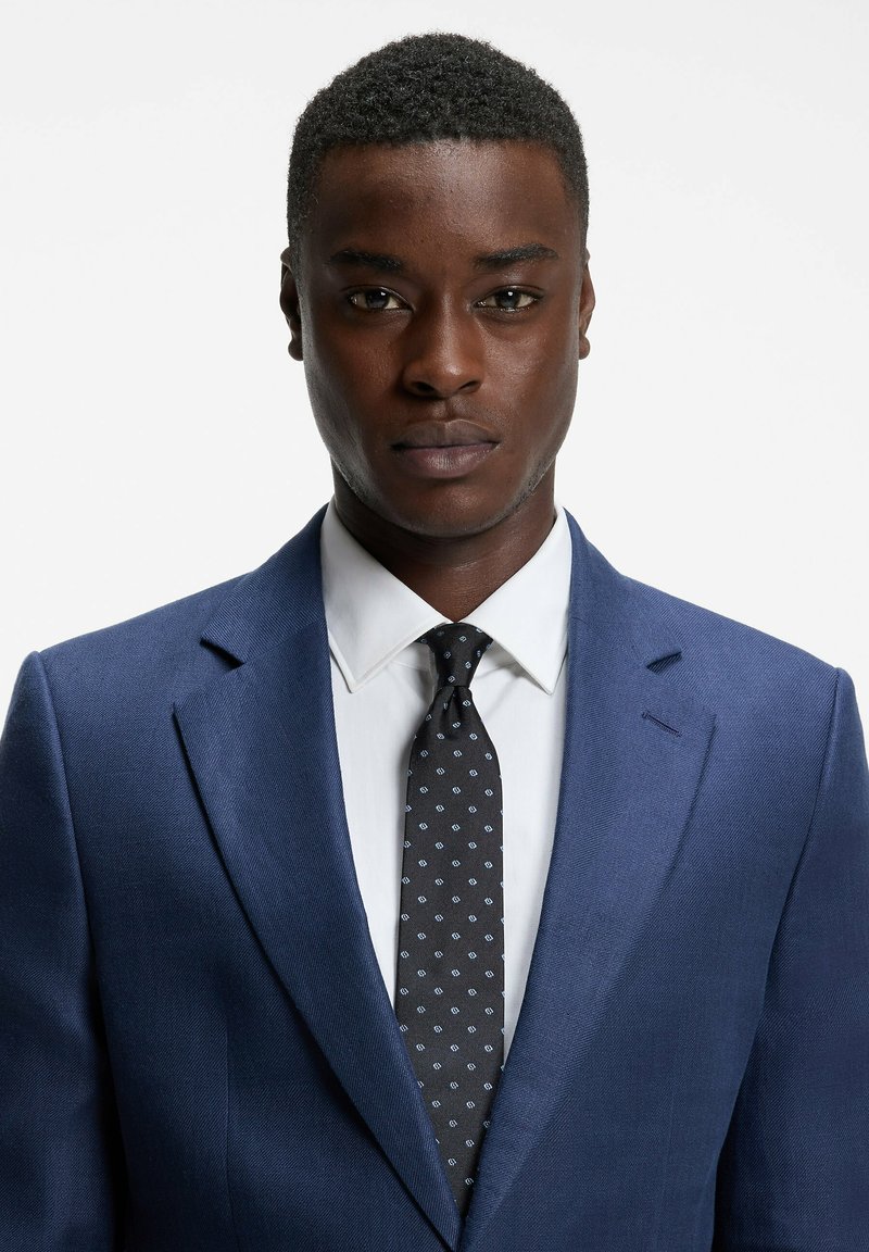 Jeune homme noir portant une veste de costume bleue, une chemise blanche et une cravate noire à motifs, faisant face à un fond blanc uni.