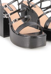 Sandalias de plataforma negras de tacón alto que presentan un material sintético brillante, múltiples tiras delgadas y acentos de hebilla en tono plateado.