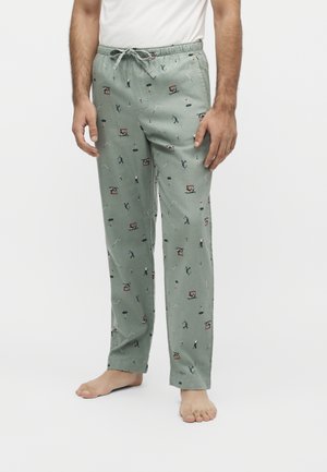Homme portant un pantalon de pyjama vert clair avec des motifs sur le thème du golf incluant des golfeurs, des voiturettes de golf et des drapeaux, associé à une chemise blanche et pieds nus.