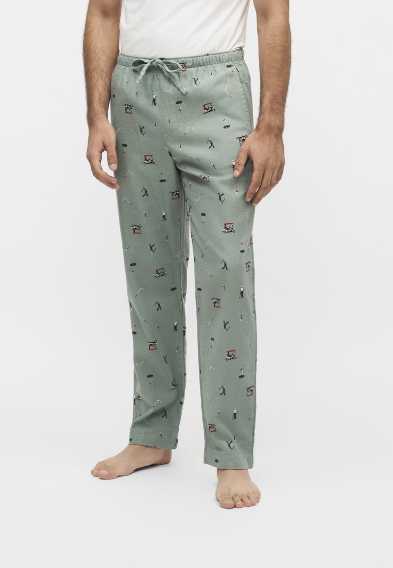Homme portant un pantalon de pyjama vert clair avec des motifs sur le thème du golf incluant des golfeurs, des voiturettes de golf et des drapeaux, associé à une chemise blanche et pieds nus.