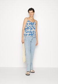 Top de corpo azul com flores brancas de lírios, alças finas, combinado com jeans azul claro e sandálias brancas. Modelo segura uma bolsa tecida bege.