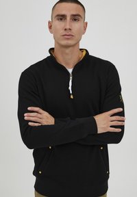 Felpa nera con zip, colletto interno giallo, polsini elasticizzati e tasche laterali con dettagli dorati. Tessuto morbido, design aderente.