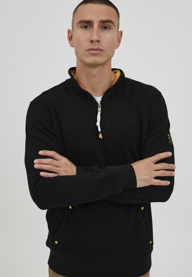 Felpa nera con zip, colletto interno giallo, polsini elasticizzati e tasche laterali con dettagli dorati. Tessuto morbido, design aderente.