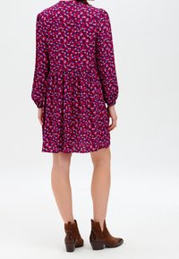Krótka fioletowa sukienka z falbanami, ozdobiona wzorem w serca w różowych i czerwonych odcieniach. Długie rękawy i klasyczny kołnierzyk. Brązowe botki za kostkę.