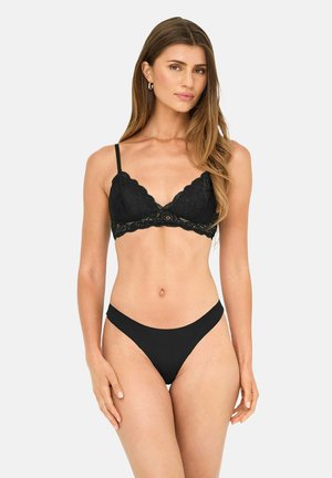 Femme aux longs cheveux portant un bralette en dentelle noire et un sous-vêtement noir taille haute assorti, debout devant un fond clair uni.