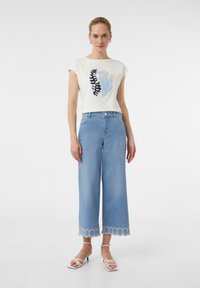 Weißes Baumwoll-T-Shirt mit floraler Stickerei, kombiniert mit hellblauen Palazzo-Jeans mit gewelltem Saum und weißen Riemchensandalen.