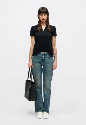 Sieviete ar garām tumšām matēm, tērpusies melnā polo krekla, zilās džinsās un baltās sporta kurpēs, kreisajā rokā tur melnu somu, stāvot pret baltu fona.