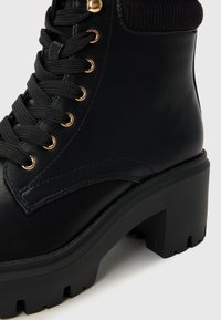 Bottine noire à talon épais, avec des œillets dorés, des lacets noirs et un col en tricot texturé. Semelle durable avec des crans pour une meilleure adhérence.