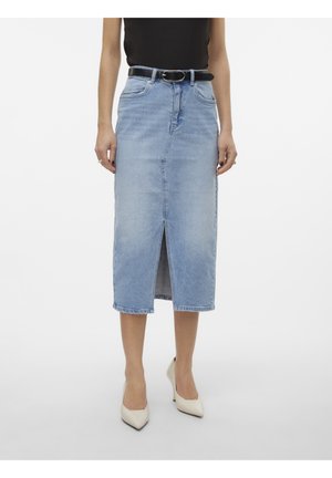 Femme portant une jupe en jean bleu clair longueur genoux avec une fente à l'avant, ceinture noire, haut noir et talons blancs pointus.