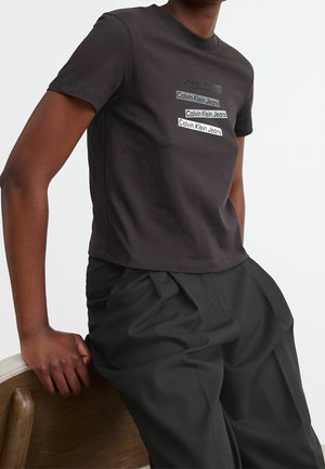 Persoon leunend op een houten stoel, draagt een zwart Calvin Klein Jeans t-shirt met gestapelde logotekst en losse zwarte broek.