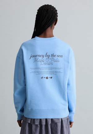 Person mit geflochtenem Haar, die einen hellblauen Sweatshirt mit dem Schriftzug „journey by the sea Marc O'Polo Denim“ auf dem Rücken trägt, kombiniert mit einem karierten Rock.