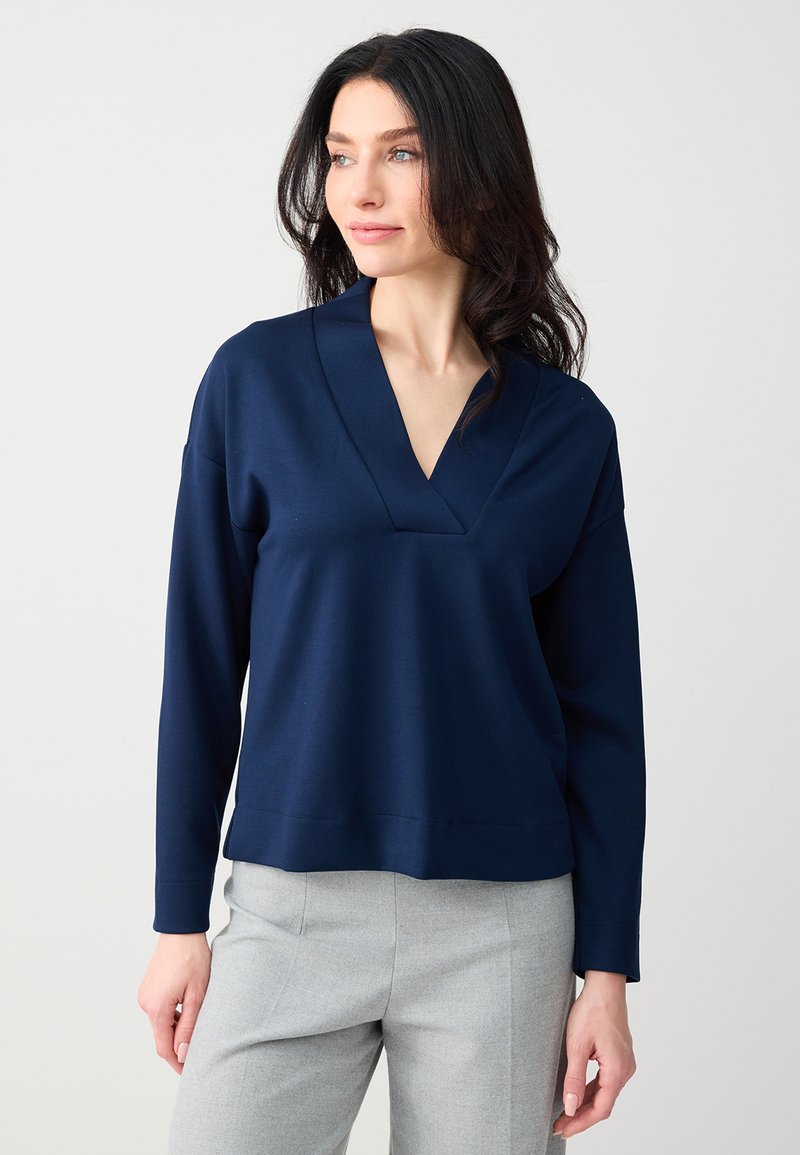 Femme aux longs cheveux bruns portant un haut bleu marine à col en V et manches longues, ainsi qu'un pantalon gris clair, regardant légèrement sur le côté.