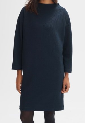 Femme portant une robe texturée bleu marine foncé, à manches longues, longueur genou, avec un col rond et des collants noirs.