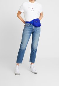 Persona con camiseta blanca de Tommy Jeans, jeans azul claro, zapatillas de deporte altas blancas y una riñonera azul, frente a un fondo liso.