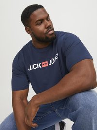 Camiseta de manga corta azul marino hecha de algodón, con un logo blanco y rojo "JACK & JONES" en el pecho, combinada con jeans de mezclilla.