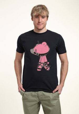 Ung mand med blondt hår iført en sort t-shirt med en pink tegneserie bjørn i seletøj og mundkurv, kombineret med lysegrønne bukser.