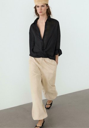 Frau geht drinnen, trägt ein schwarzes Hemd mit Knöpfen, beige weite Hose, schwarze Sandalen und einen geflochtenen Hut, Hände in den Taschen.