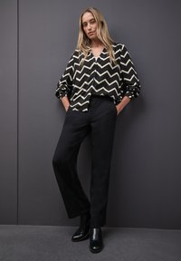 Schwarz-weiß gemusterte Bluse mit Zickzack-Muster und Knopfleiste, kombiniert mit hochgeschnittenen schwarzen Hosen und glänzenden schwarzen Knöchelstiefeln.