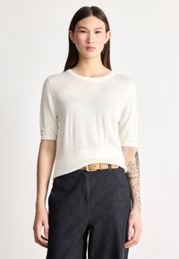 Lahka kremna pulover s kratkimi rokavi in okroglim izrezom. Oblečen s temnimi denim hlačami in pasom iz rjavega usnja. Gladek videz, minimalističen dizajn.