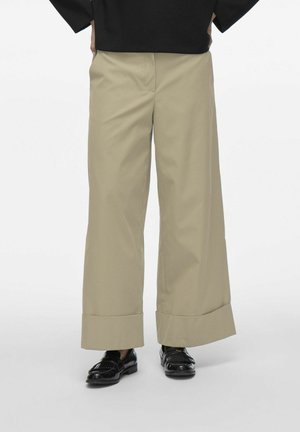 Pantalones beige de pierna ancha con una textura suave, con un dobladillo doblado. Combinados con mocasines negros de charol, que presentan un acabado brillante.