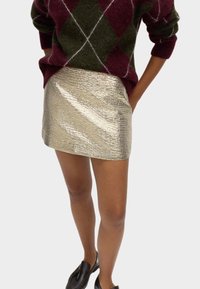 Jersey de lana con un patrón de rombos en burdeos y verde, combinado con una falda mini dorada brillante y texturizada y mocasines negros.