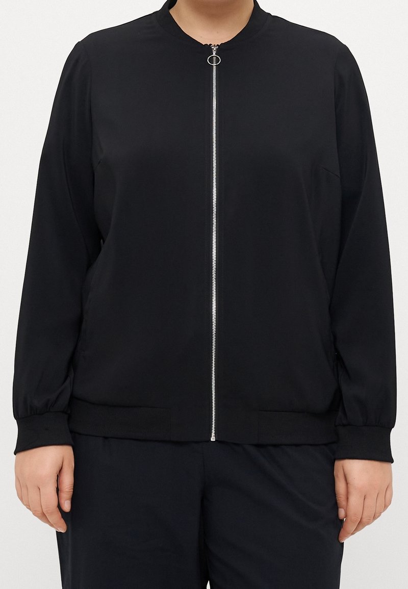 Personne portant une veste noire zippée à manches longues et col rond, debout devant un fond uni clair.