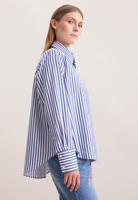 Vrouw met steil haar draagt een oversized blauw-wit gestreept shirt met verticale strepen en lichtblauwe jeans, staand tegen een effen lichte achtergrond.