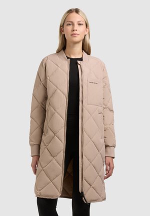 Donna con un lungo giubbotto trapuntato beige indossato sopra abiti neri, in piedi con un'espressione neutra contro uno sfondo chiaro e semplice.