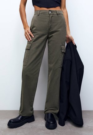 Pantalons cargo vert olive avec poches latérales, une coupe droite et un tissu robuste. Associés à des bottes noires chunky et une veste noire.