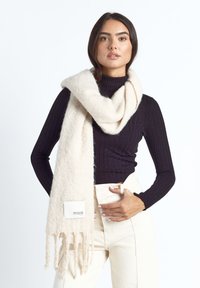 Kobieta w czarnym prążkowanym golfie, kremowych spodniach z wysokim stanem oraz w grubym kremowym szaliku z frędzlami i metką marki Maze.