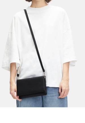 Persona que lleva una camisa blanca suelta y jeans azules, llevando un bolso cruzado de cuero negro con una correa delgada para el hombro.