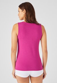 Mujer con cabello castaño que lleva una blusa sin mangas magenta y pantalones cortos blancos, de pie y mirando hacia afuera contra un fondo gris claro liso.