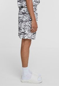 Shorts blancs avec motifs abstraits noirs, taille élastique et poches latérales. Portés avec des chaussettes blanches et des sandales slide, offrant une coupe décontractée.