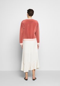 Sudadera rosa con puños acanalados combinada con una falda midi blanca a rayas con un dobladillo con volantes. Sandalias sencillas completan el conjunto.