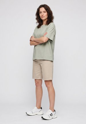 Woman with curly brown hair stands with arms crossed wearing light green t-shirt, beige shorts, and white sneakers with black heart design.