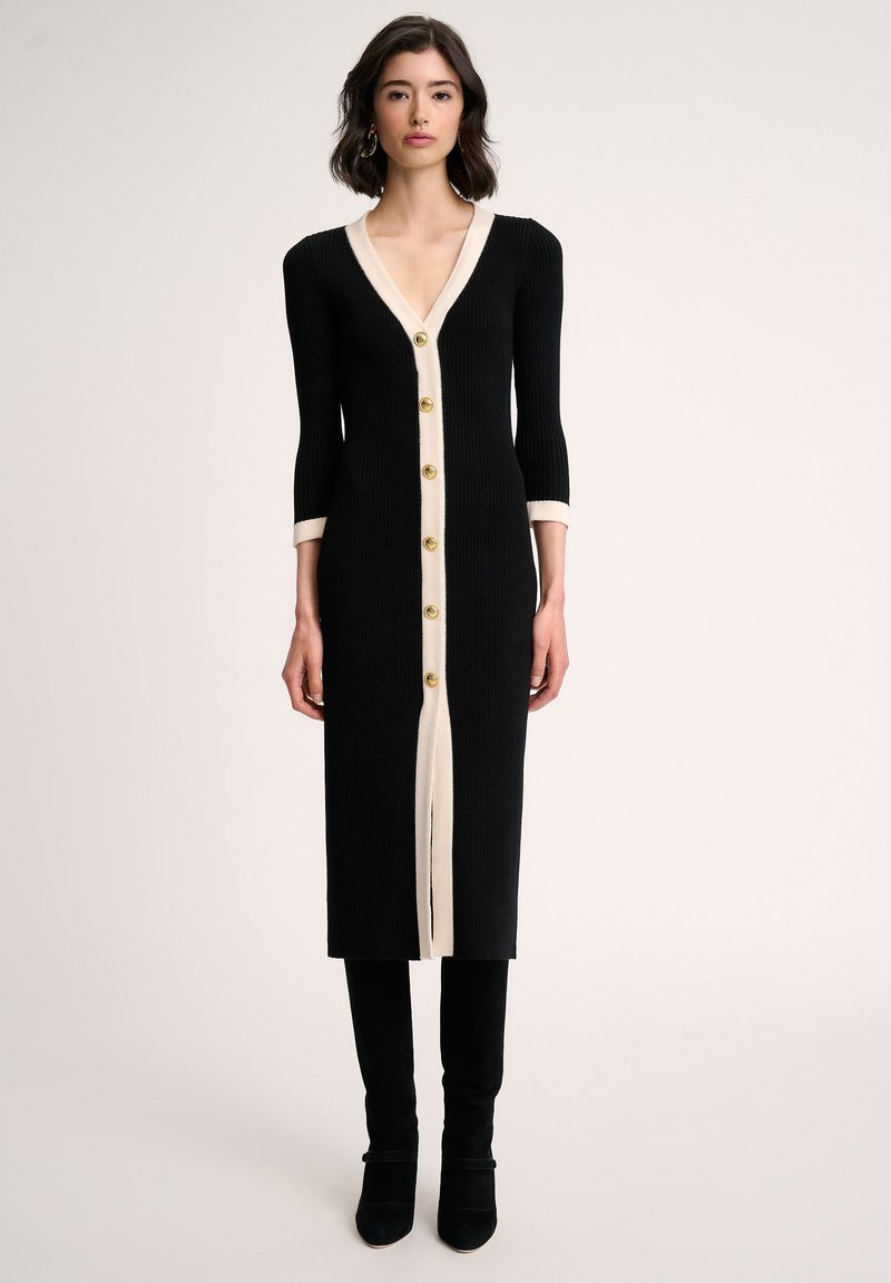 Long cardigan noir côtelé avec des bordures crème et six boutons dorés, présentant un décolleté en V et des manches trois-quarts. Texture de tissu lisse.