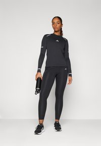 Schwarzes, langärmliges Sportoberteil mit reflektierenden Akzenten, kombiniert mit schwarzen Leggings und schwarzen Sportschuhen. Wasserflasche in einer Hand gehalten.