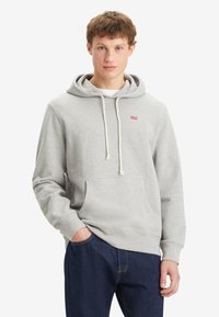 Sudadera gris con capucha ajustable, bolsillo frontal y logotipo rojo en el pecho. Combinada con jeans azul oscuro, de corte relajado y textura suave.
