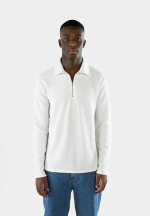 Långärmad vit tröja med dragkedja i halsen, strukturerad tyg och avslappnad passform; matchad med blå denimjeans.