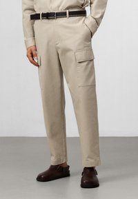 Pantaloni cargo beige con tasche laterali, design a gamba dritta e una lieve textura. Indossati con una cintura nera e scarpe marrone scuro.