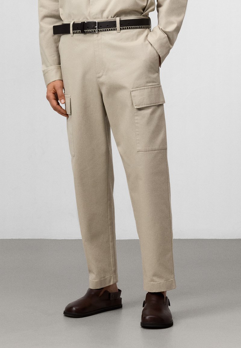 Beige cargobroeken met zijzakken, recht-lijn ontwerp en een subtiele textuur. Gecombineerd met een zwarte riem en donkerbruine schoenen.