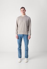 Lys grå hettegenser med ribbestrikket halslinning og mansjetter, parret med lyseblå denimjeans. Modellen står foran en nøytral bakgrunn.