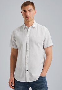 Camisa blanca de manga corta con botones, hecha de un tejido ligero. Presenta un logo sutil en el pecho y un dobladillo curvado. Se lleva con unos vaqueros azules.