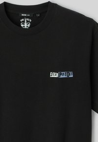 T-shirt en coton noir à coupe ample, arborant un petit logo graphique en texte blanc et bleu "Sex Pistols" sur la poitrine.